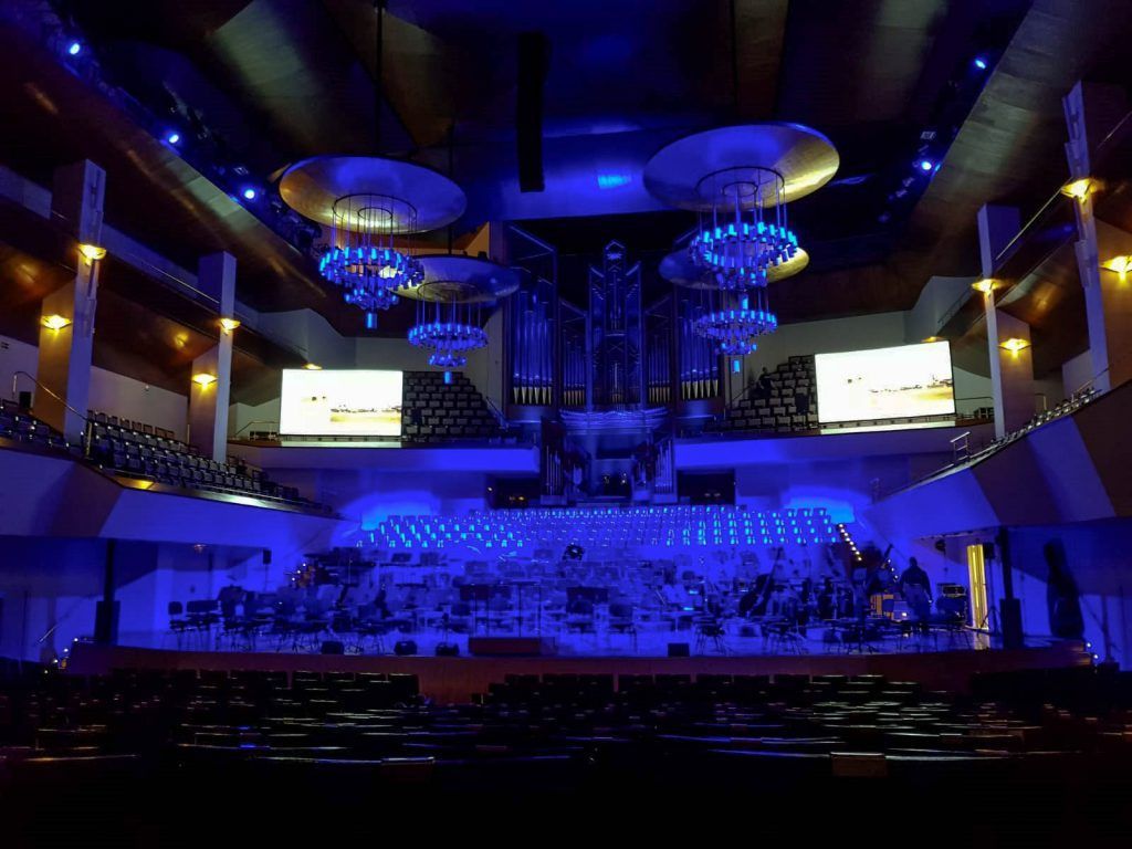 Teatro o sala de conciertos con iluminación azul, candelabros, escenario y butacas orientadas hacia una gran pantalla.