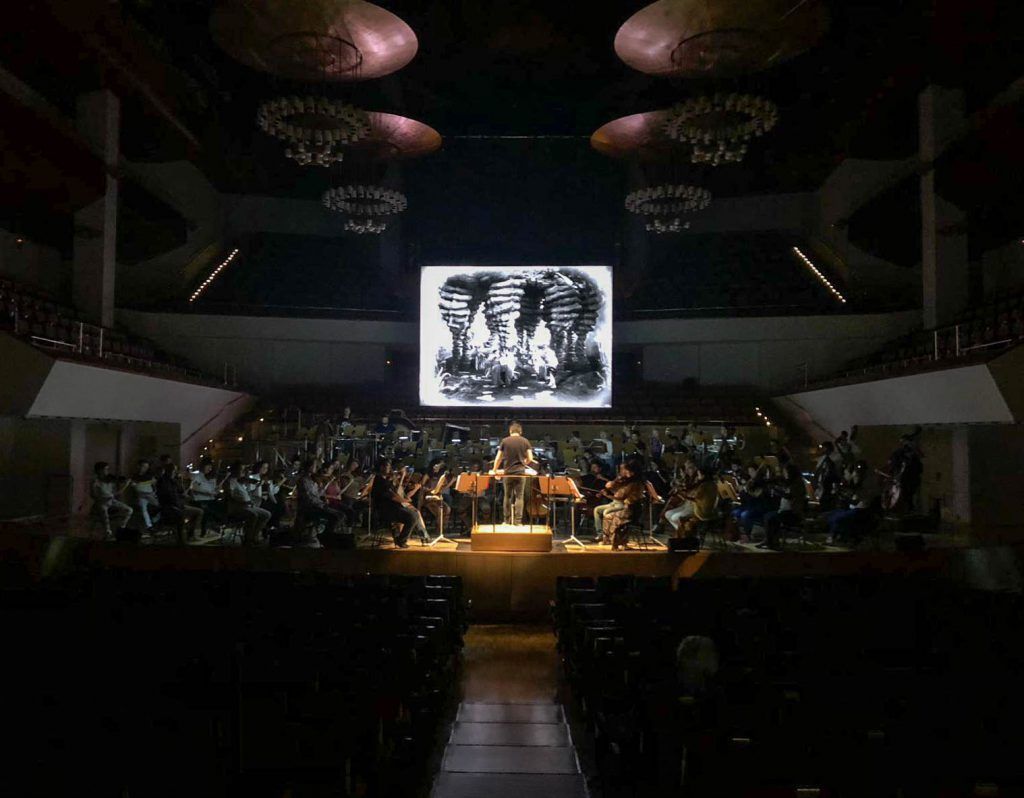 Orquesta actuando en un escenario con poca luz y una gran pantalla proyectada sobre ellos.