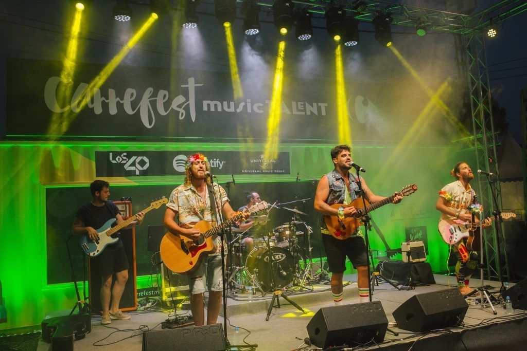 Banda actuando en un escenario iluminado con luz verde, con guitarras, batería y focos amarillos brillantes.