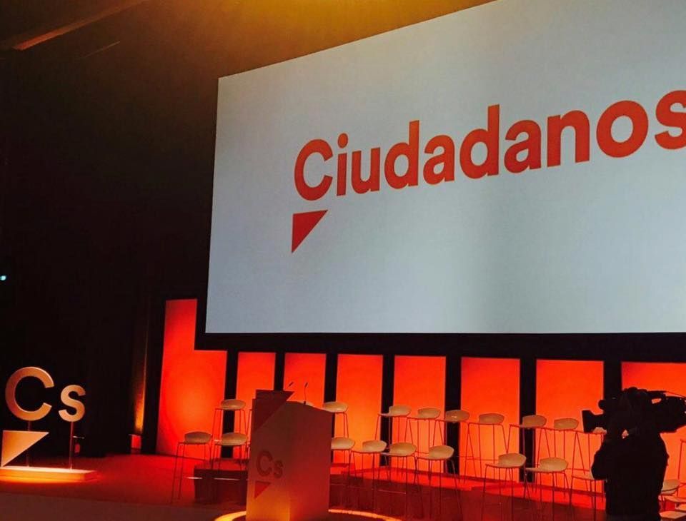 Escenario con iluminación roja, podios, sillas y una gran pantalla que muestra “Ciudadanos”.