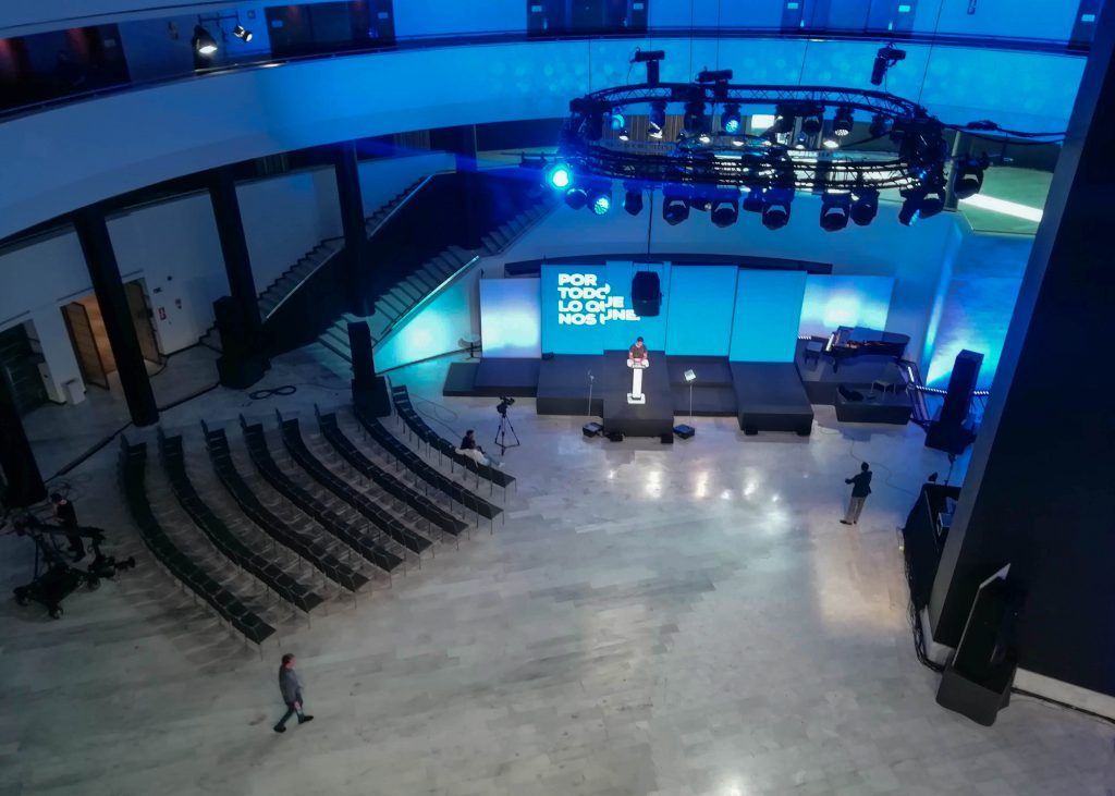 Vista panorámica de un escenario de auditorio vacío con iluminación azul, filas de asientos y una gran pantalla de fondo.