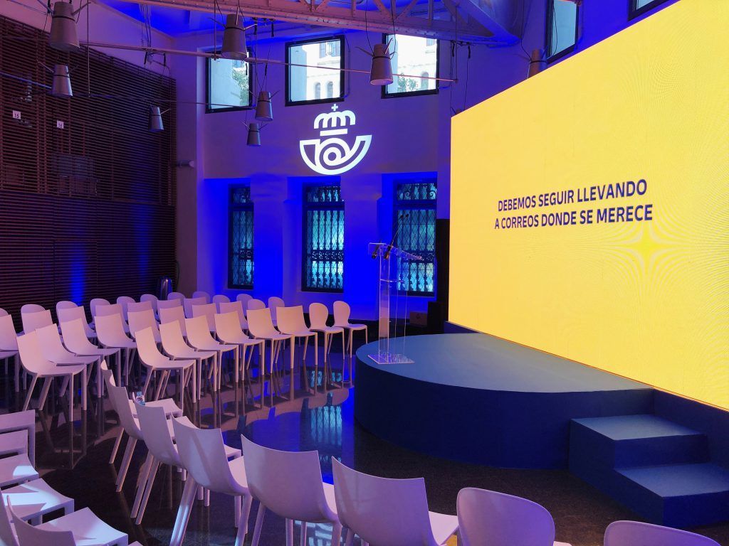 Escenario para conferencias con filas de sillas, iluminación azul y una gran pantalla de presentación amarilla.