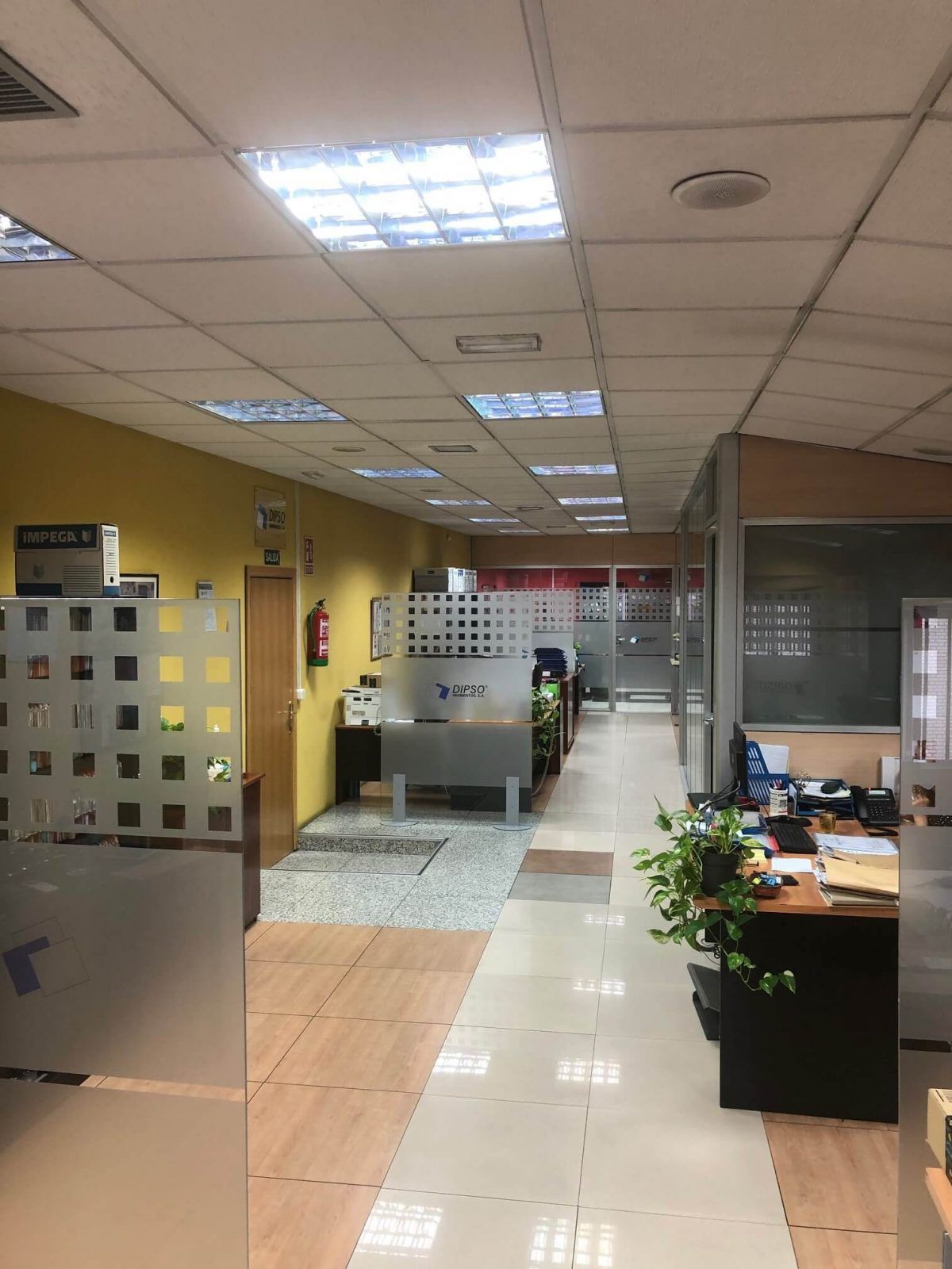Pasillo de oficina con cubículos, escritorios y paredes de vidrio esmerilado; iluminación fluorescente.