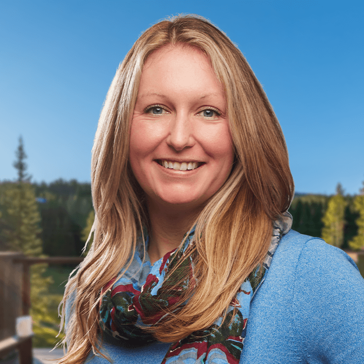 Woman with blonde hair smiling, wearing a blue sweater and scarf, with a wooded background.