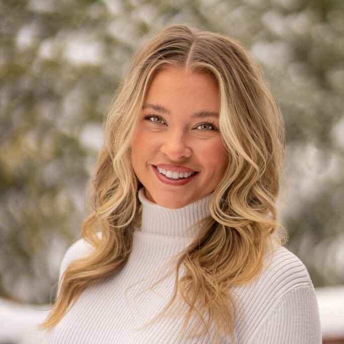 Woman with blonde wavy hair smiles, wearing a white turtleneck sweater, set against a snowy backdrop.