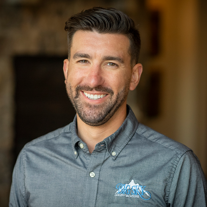 Man with dark hair and beard, smiling, wearing gray button-down shirt with mountain logo.