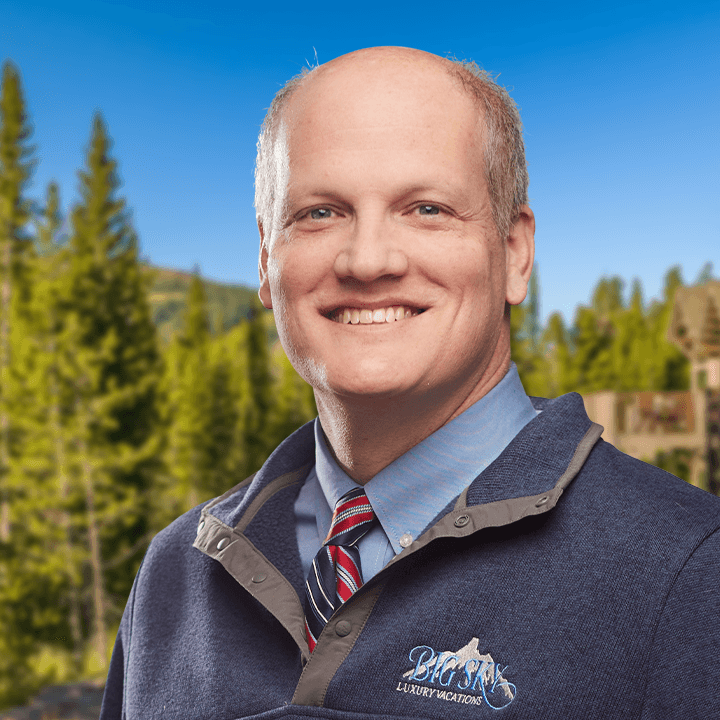 Man smiling, wearing a blue sweater and tie, with a mountain logo; background with trees and blue sky.