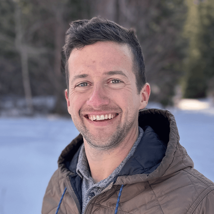 Man smiles at the camera in front of a snowy background, wearing a brown jacket.