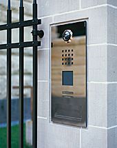 Eine Türklingel aus Edelstahl ist an einer Backsteinmauer neben einem Zaun montiert.