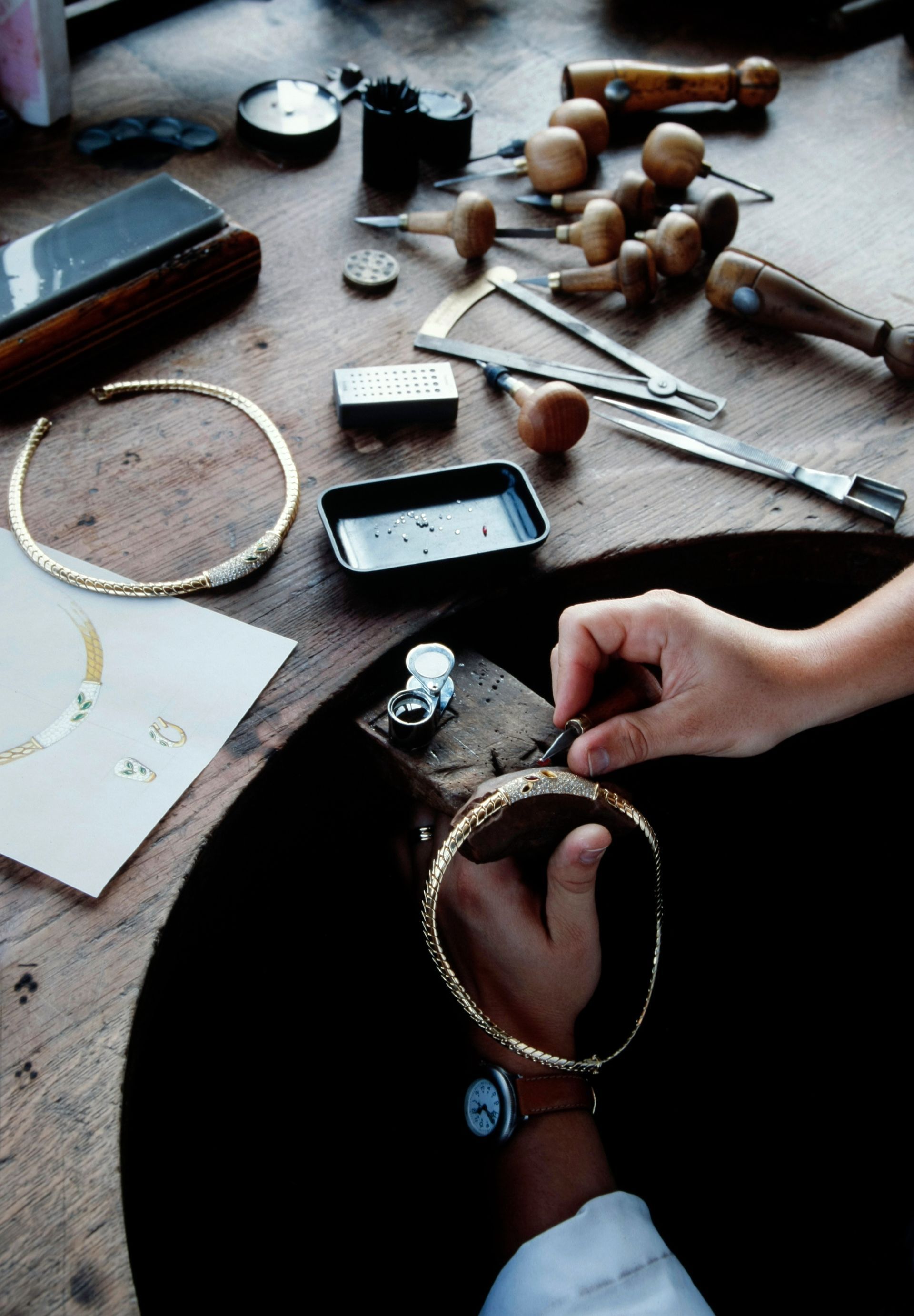 Des mains assemblent un collier en or sur un établi de bijouterie, avec des outils et des pièces finies.