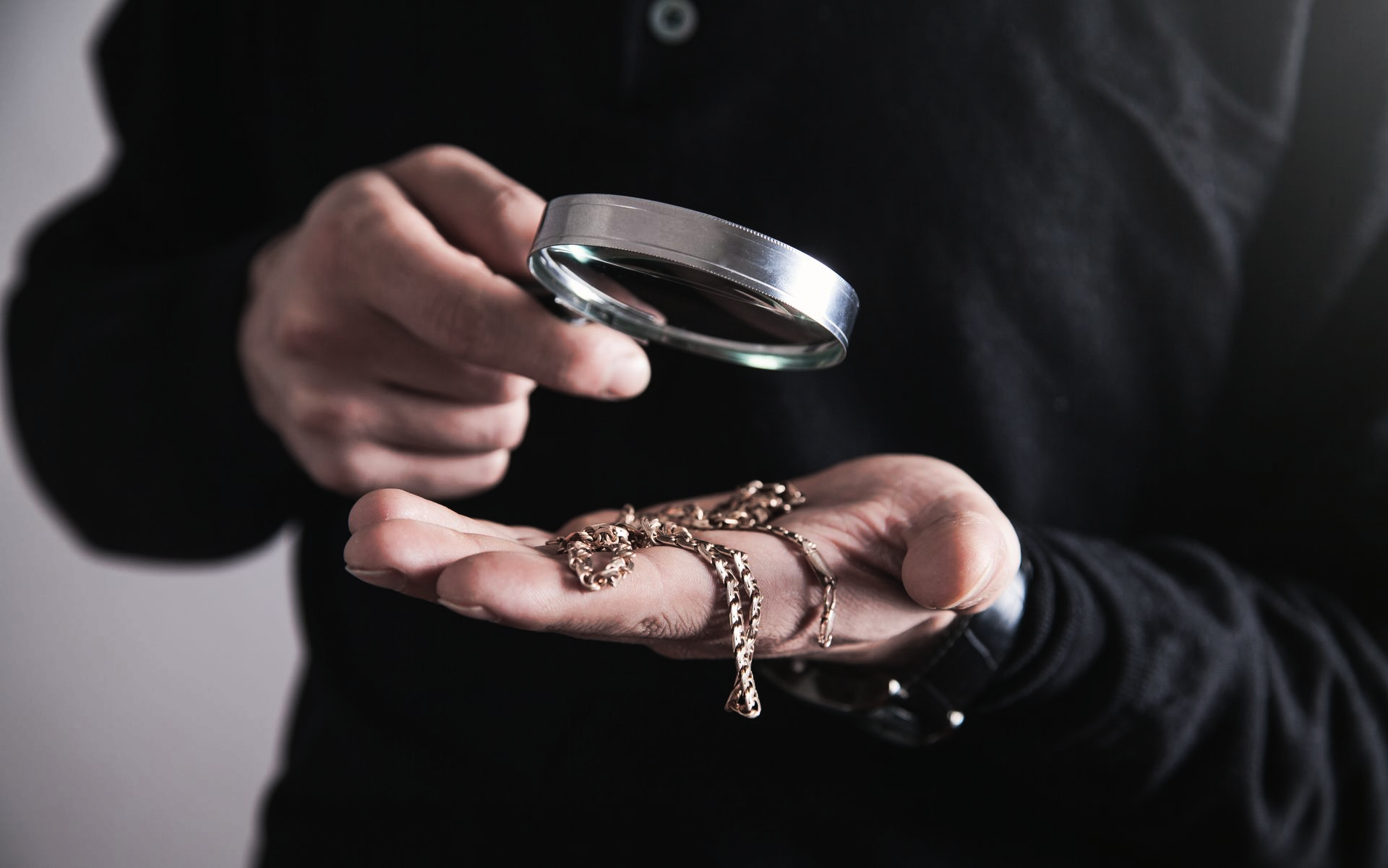 Une personne tenant une loupe au-dessus d'une main tenant des bijoux sur fond sombre