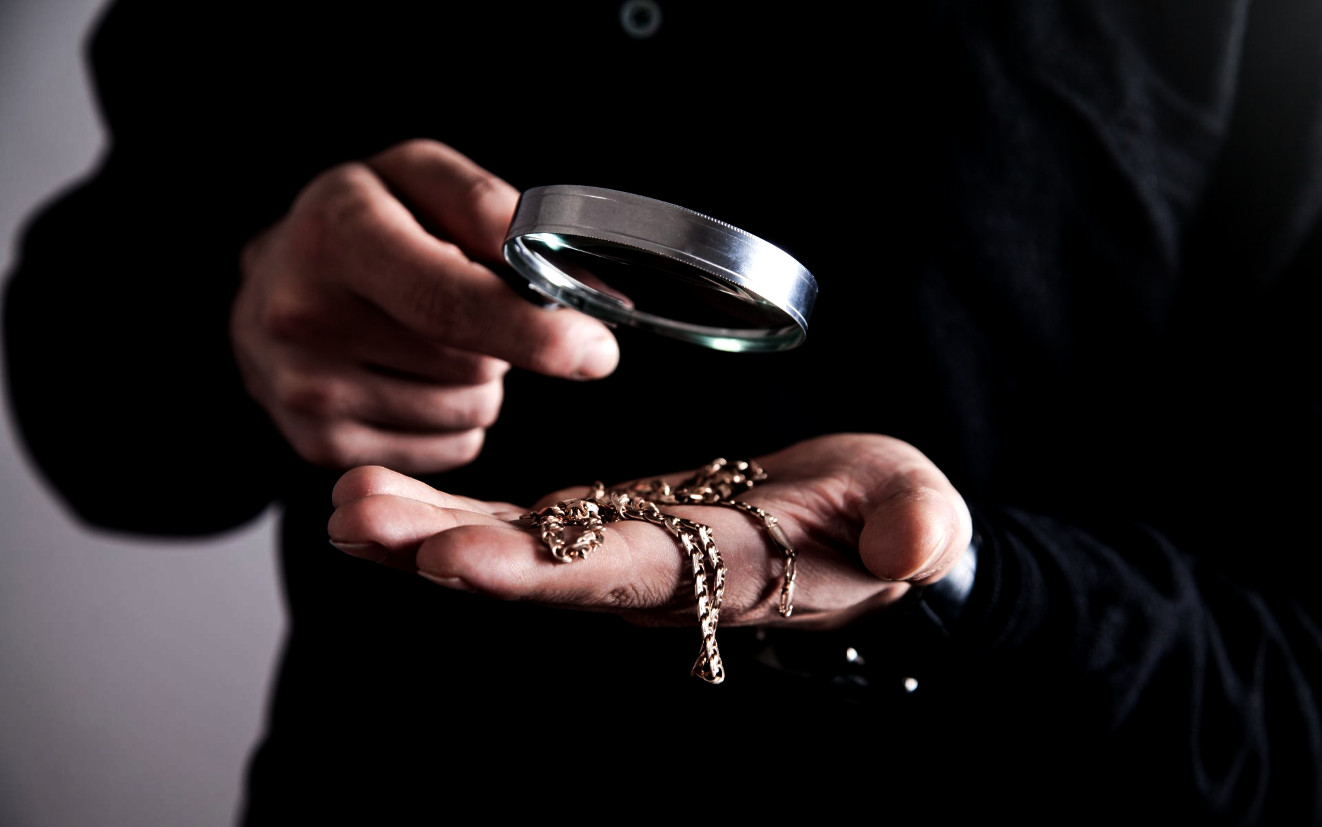 Personne tenant une loupe au-dessus d'une main portant des bijoux sur fond sombre