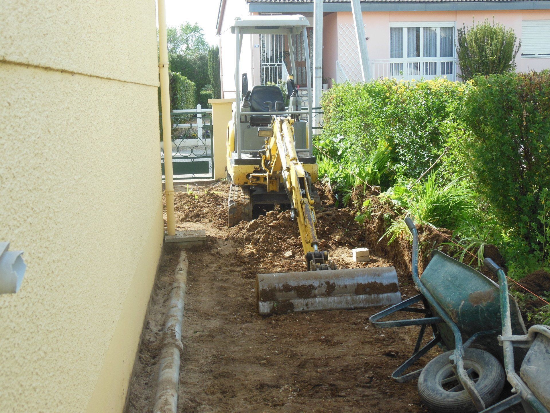 Tranchée dans le jardin avec une pelleteuse jaune à côté d'une maison et d'une haie, de la terre empilée le long du chemin.