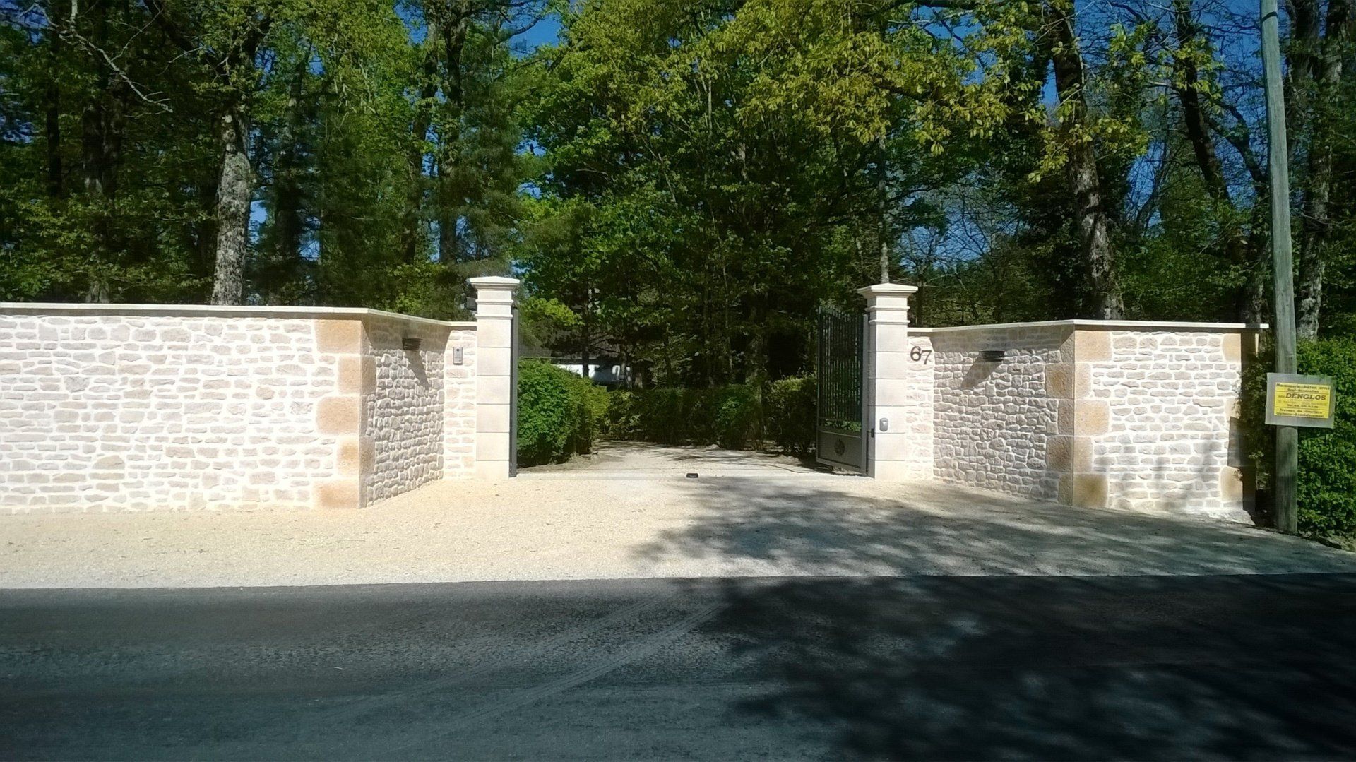Porte d'entrée et mur en pierre donnant sur une zone boisée, avec un chemin de gravier et des piliers en brique.