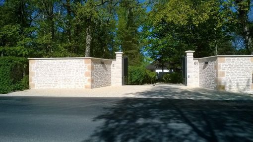 Portail d'entrée et mur en pierre avec allée ouverte, flanqué de murets et d'arbres baignés de lumière naturelle.