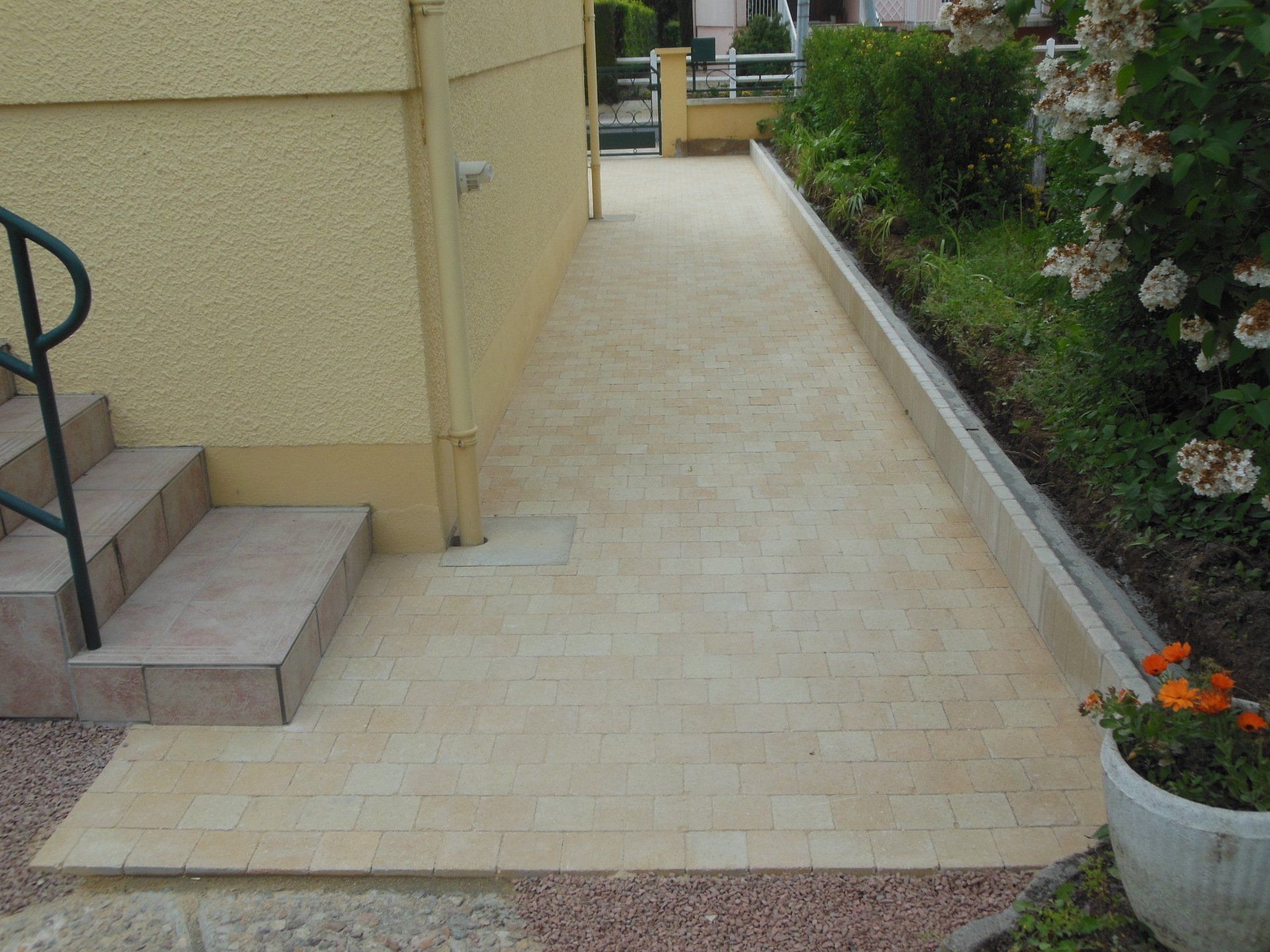 Allée étroite carrelée de beige longeant une maison, avec des marches à gauche et une jardinière à droite.
