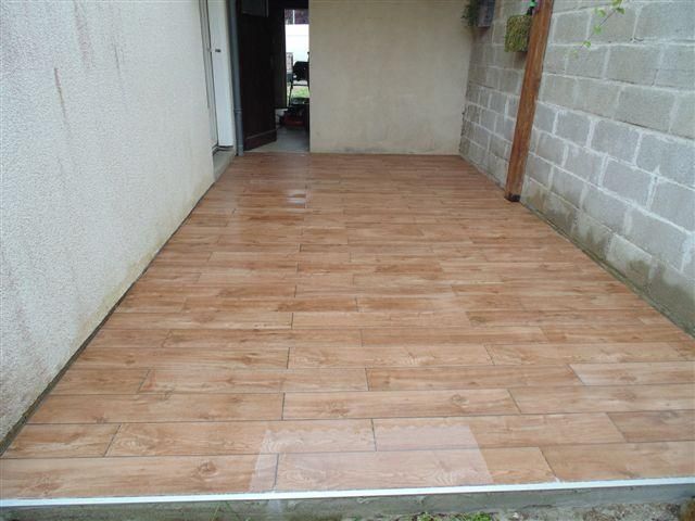 Terrasse carrelée vide, jouxtant un mur blanc et un mur en parpaings gris, vue depuis une porte.