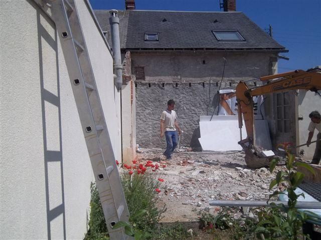 Chantier de construction avec échelle et excavatrice à côté d'un bâtiment ; un ouvrier se tient au milieu des décombres.