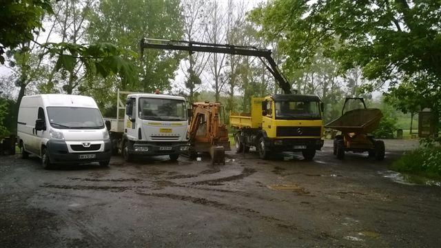 Parking avec quatre camions utilitaires sous les arbres, sur une route mouillée et boueuse.
