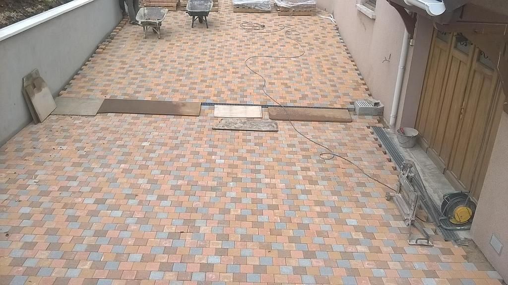 Cour pavée de carreaux emboîtables beiges et orange, bordée de murs et d'un portail en bois sur la droite.