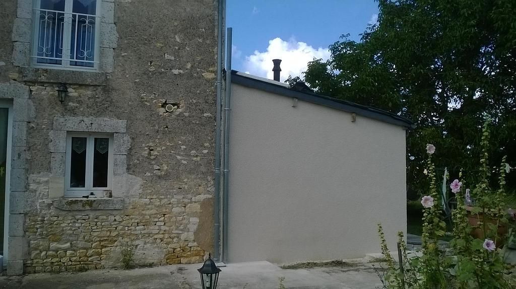 Façade en pierre d'une maison jouxtant un mur blanc et un arbre dans une cour/jardin ensoleillée