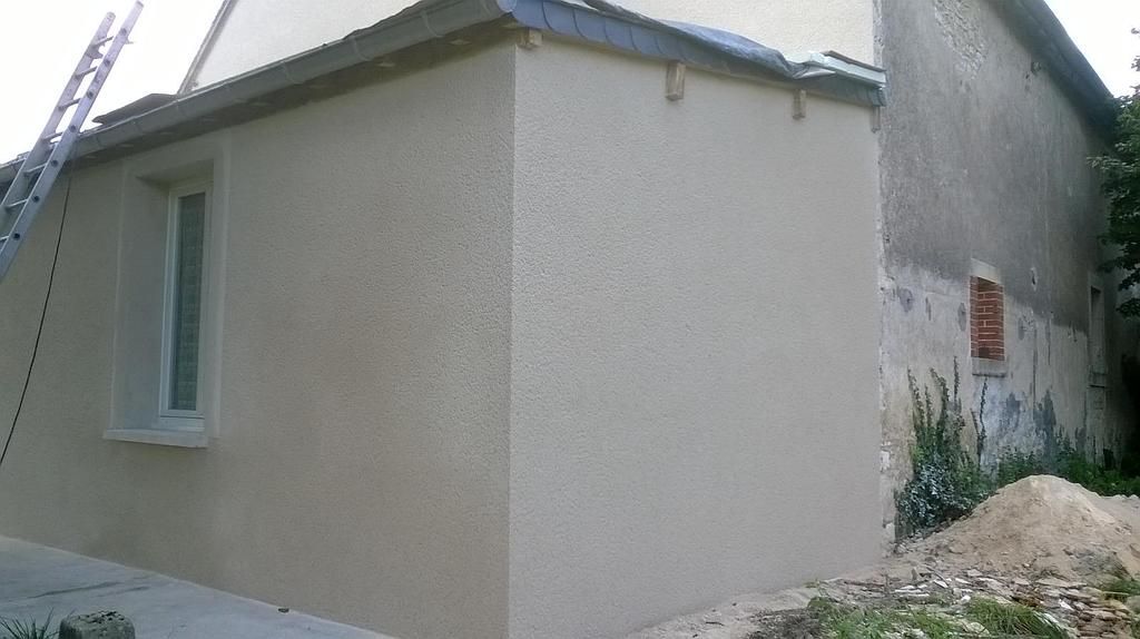 Petite maison beige avec une fenêtre et un toit en pente, située à côté d'un chemin de terre.
