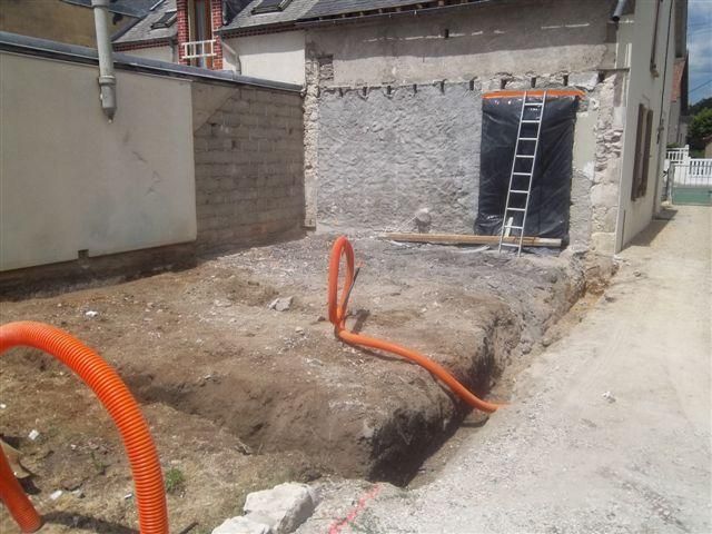 Tranchée de chantier à côté d'un bâtiment, avec des tuyaux orange et une échelle appuyée contre un mur.