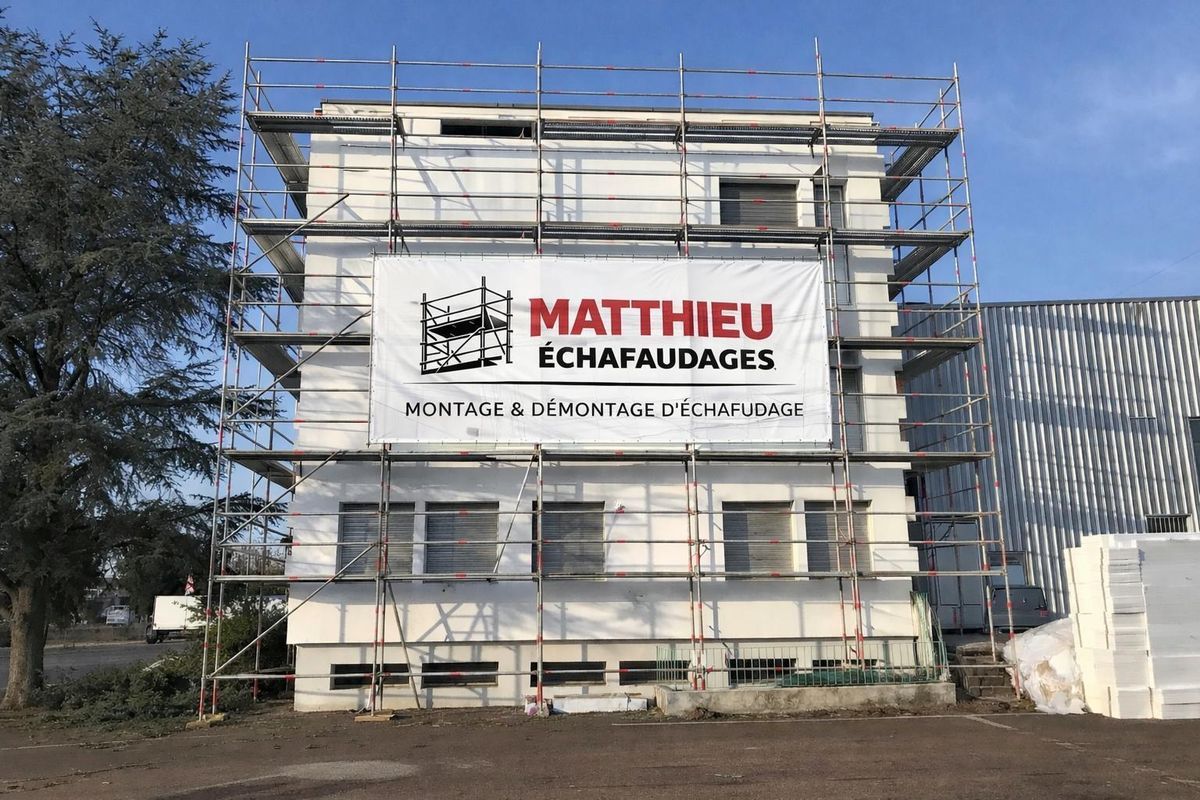 Un bâtiment blanc sous un ciel bleu, recouvert d'échafaudages métalliques avec une pancarte « Matthieu Échafaudages » affichée dessus.