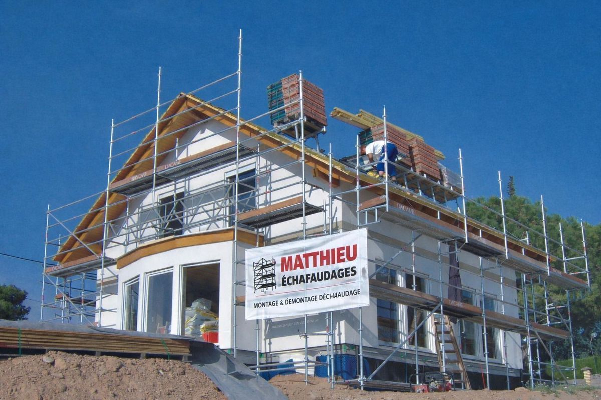 Une maison blanche en construction, entourée d'échafaudages et ornée d'une banderole publicitaire, se détache sur un ciel d'un bleu éclatant.