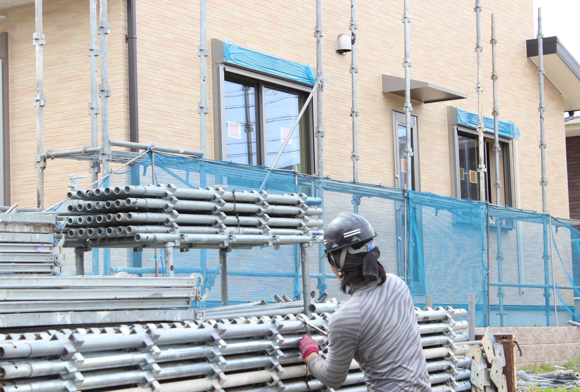Une personne portant un casque de chantier travaille avec des tubes d'échafaudage métalliques empilés devant une maison en construction.