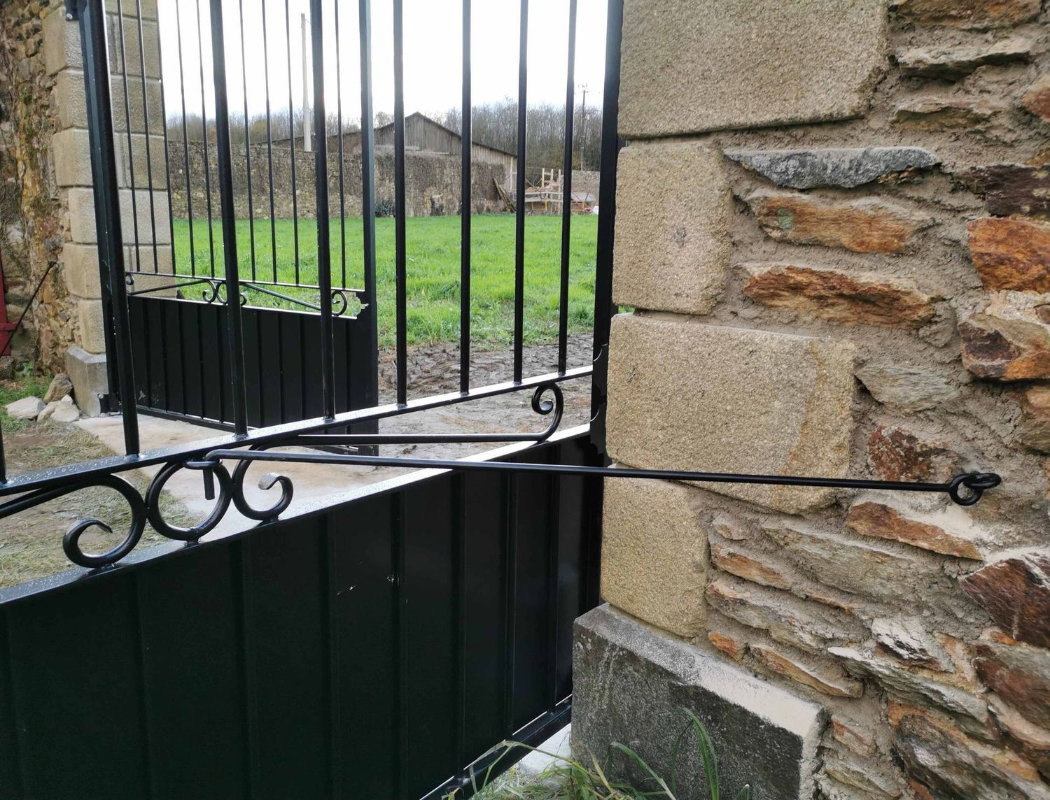 Portail en métal noir encastré dans un mur de pierre, muni d'un bras décoratif pour le maintenir ouvert.