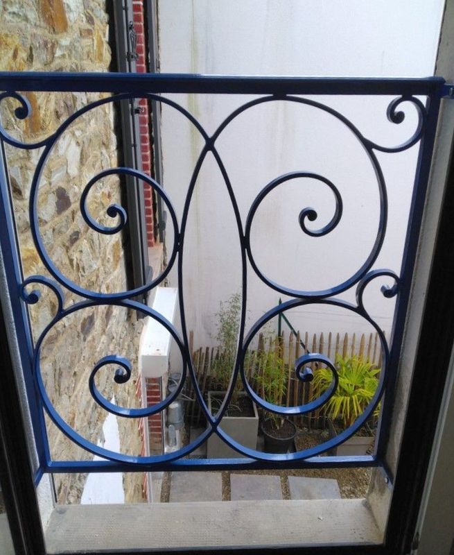 Rambarde de balcon en fer forgé bleu ornée de volutes, donnant sur un petit patio planté de plantes.
