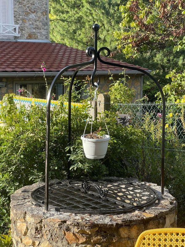 Un puits en pierre avec une structure métallique soutenant un pot de fleurs blanc suspendu. Une végétation luxuriante l'entoure.