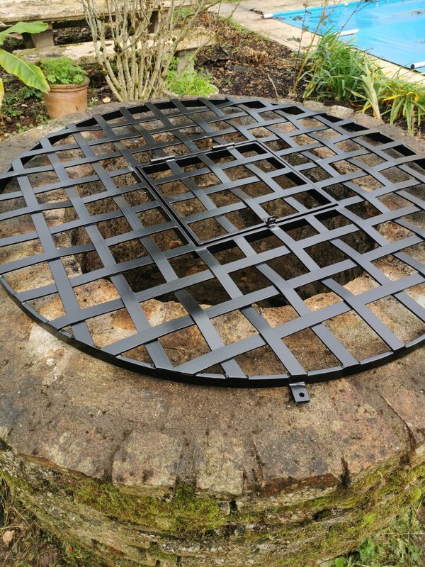 Couvercle de puits en treillis métallique noir sur un puits en briques, avec un petit panneau d'accès carré.