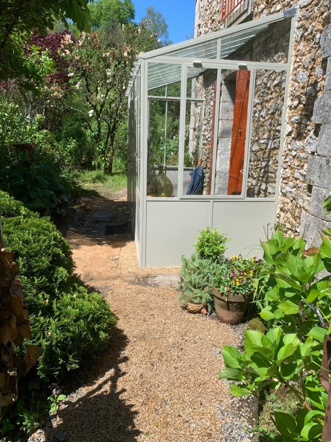 Allée de gravier menant à une serre jouxtant un bâtiment en pierre, avec des plantes au premier plan.