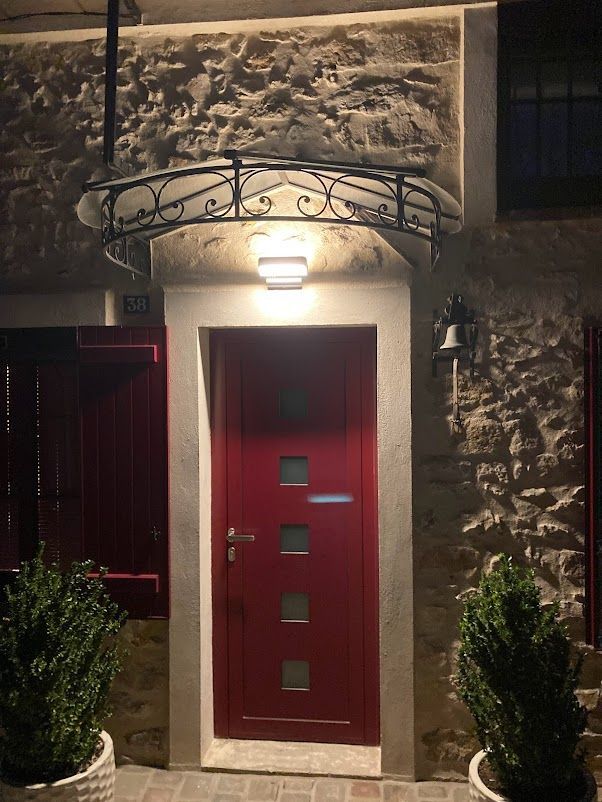 Porte rouge à carreaux de verre sous un auvent lumineux. Façade en pierre avec plantes en pot de part et d'autre.