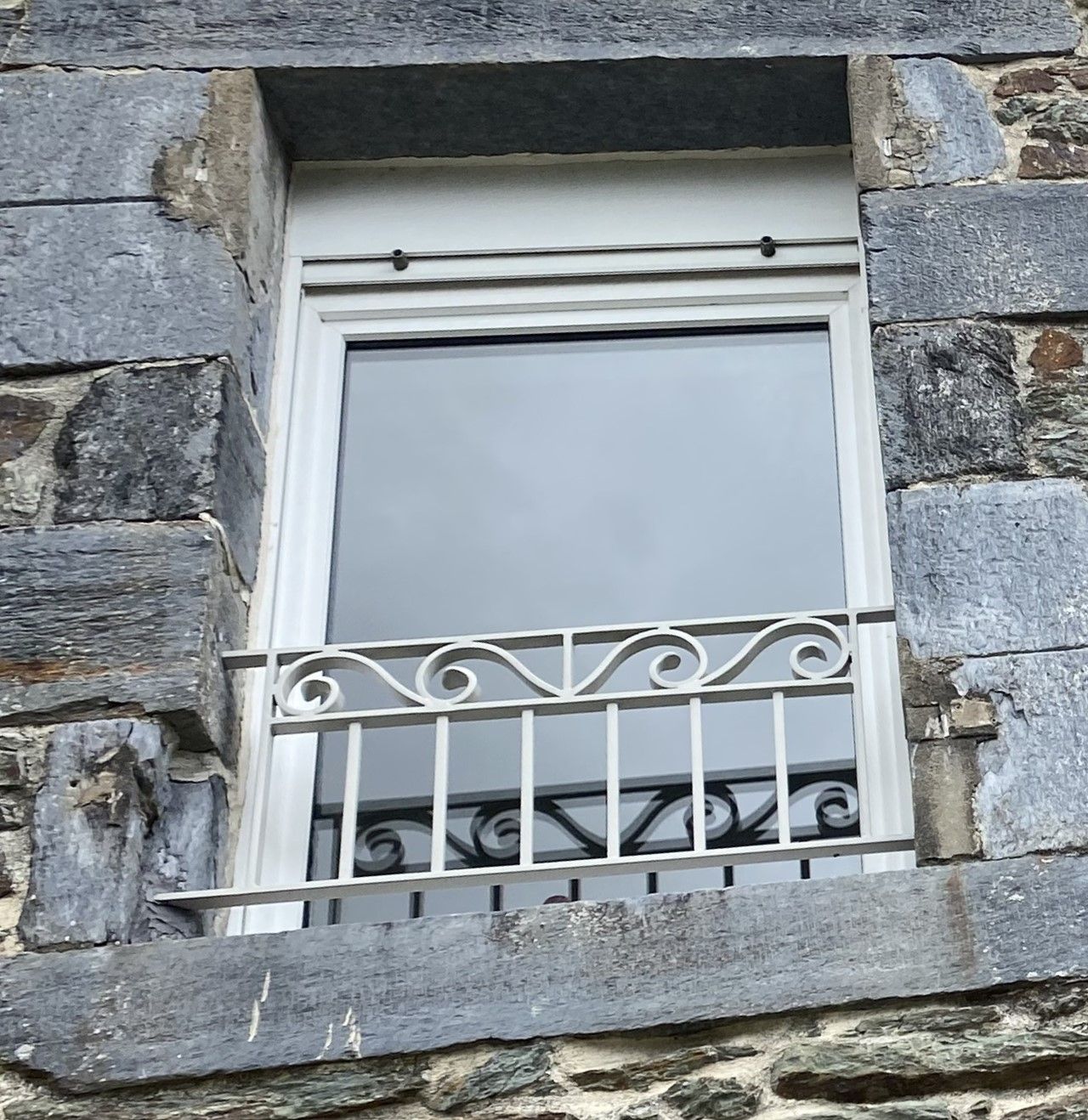 Fenêtre dans un mur de pierre, cadre blanc, barreaux de sécurité en métal ouvragé.