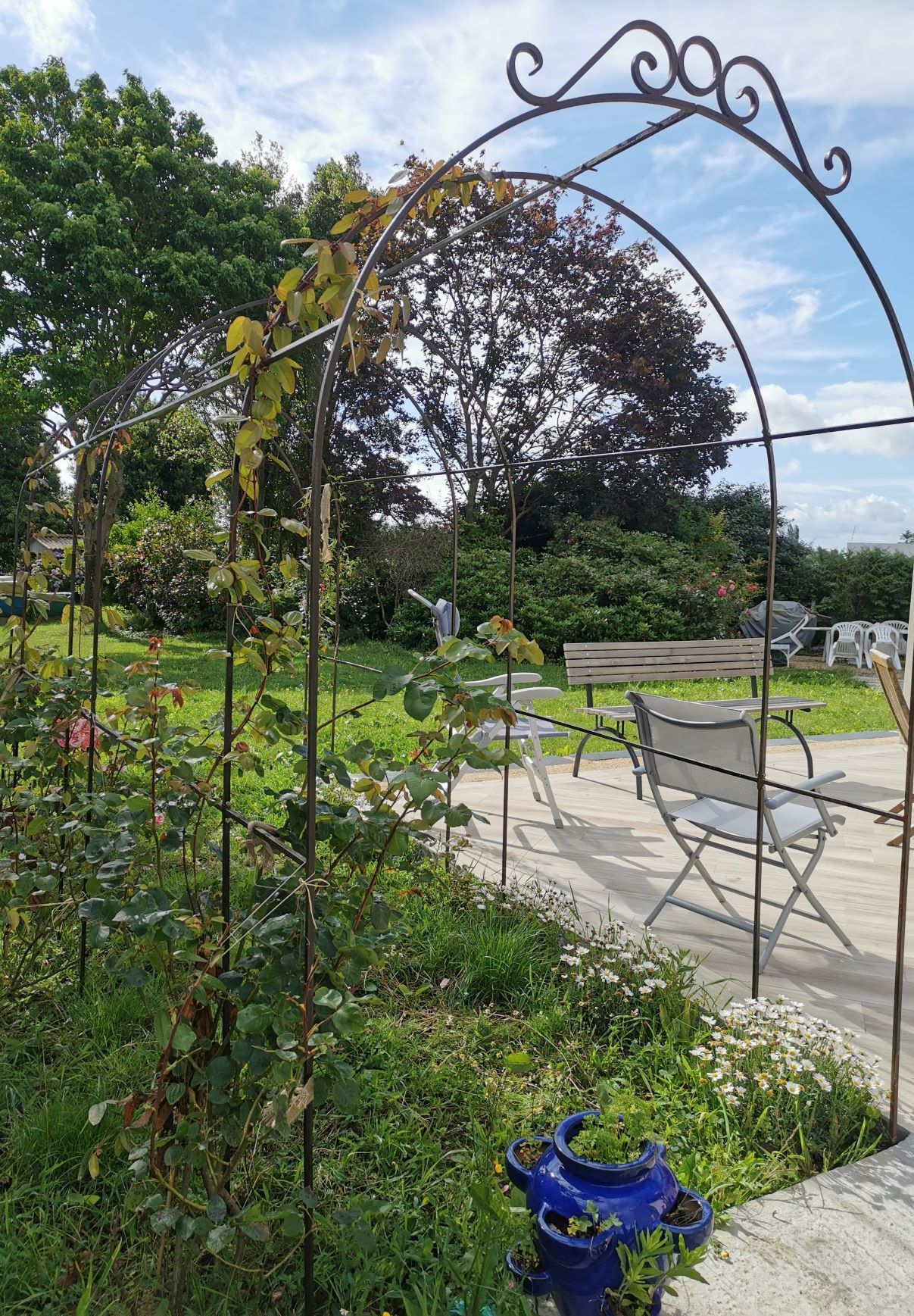 Arche de jardin avec plantes grimpantes, rosiers, pelouse, coin salon et pot bleu.