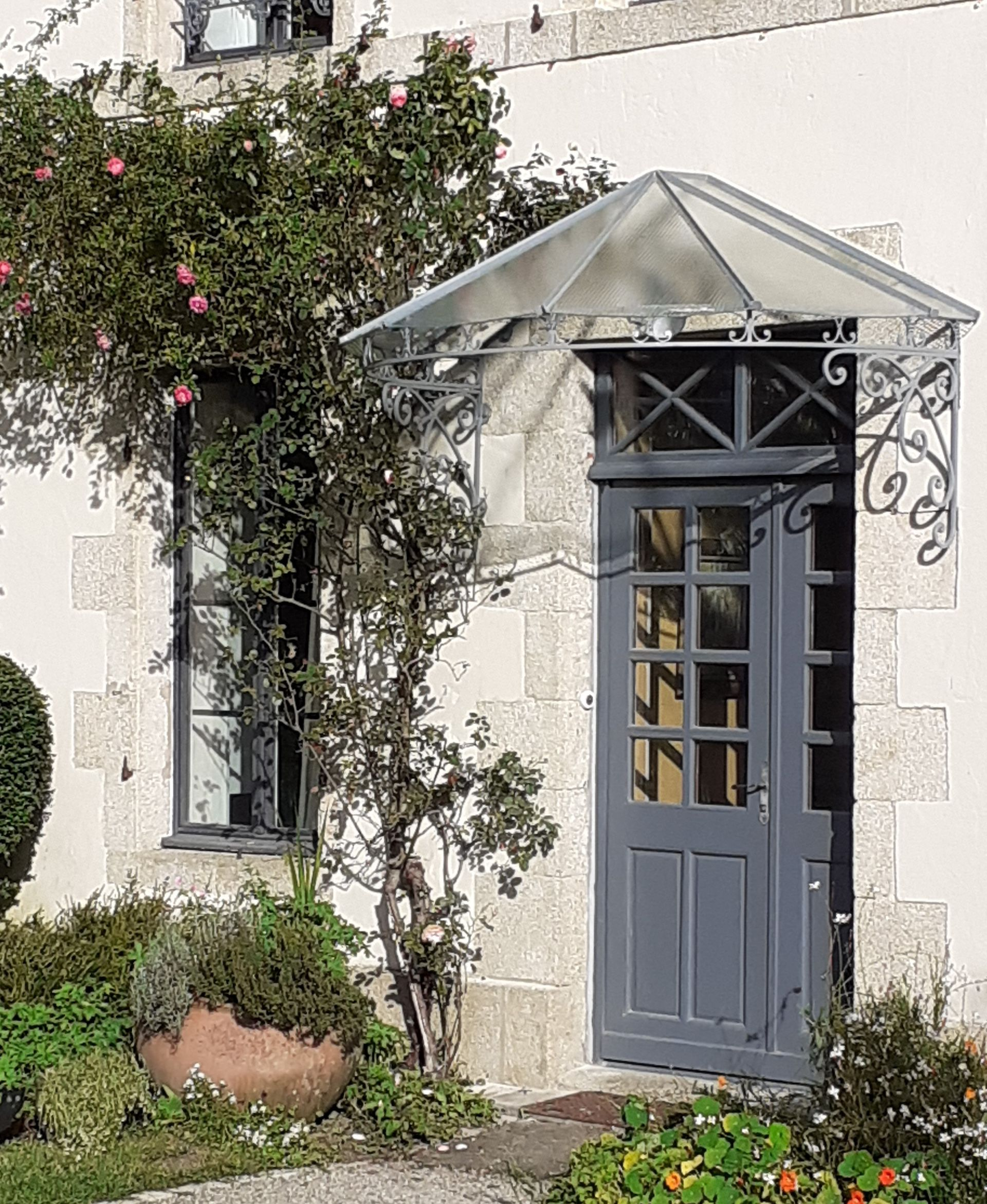 Porte grise sous un auvent en verre, avec un rosier grimpant, intégrée à la façade d'un bâtiment en pierre.