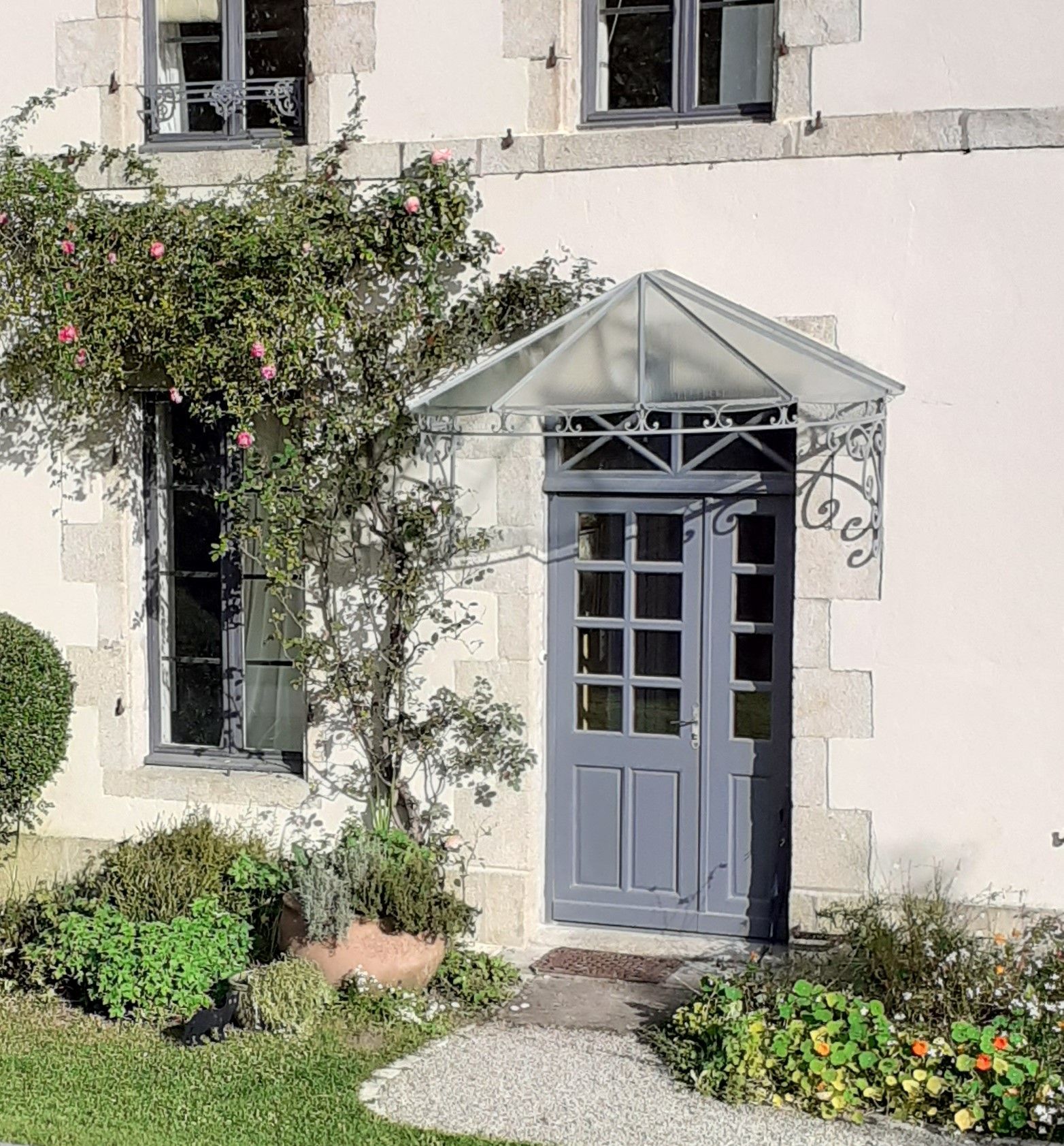 Porte grise avec auvent en verre, sur une maison en pierre avec un rosier grimpant.