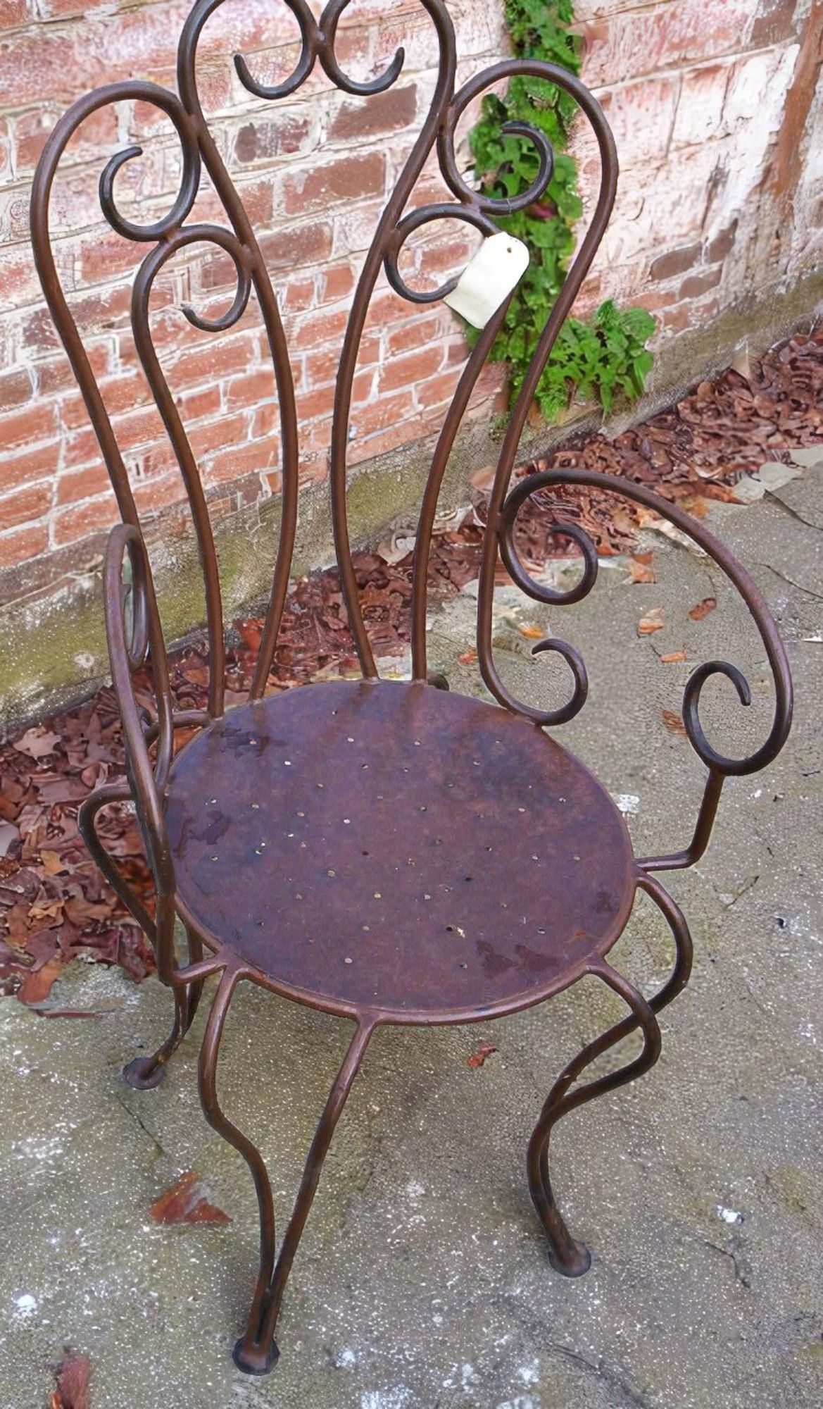 Chaise en fer forgé, à assise circulaire et dossier incurvé, adossée à un mur de briques.