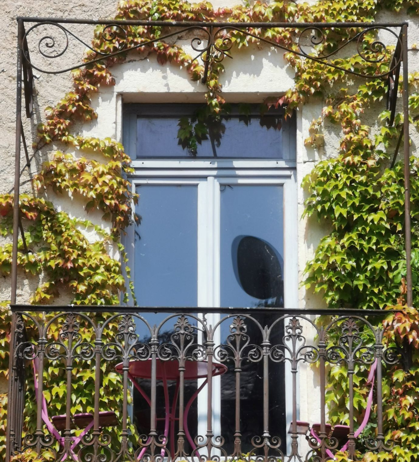 Fenêtre avec balcon en fer forgé, mur de pierre recouvert de lierre. Table et chaises roses sur le balcon.