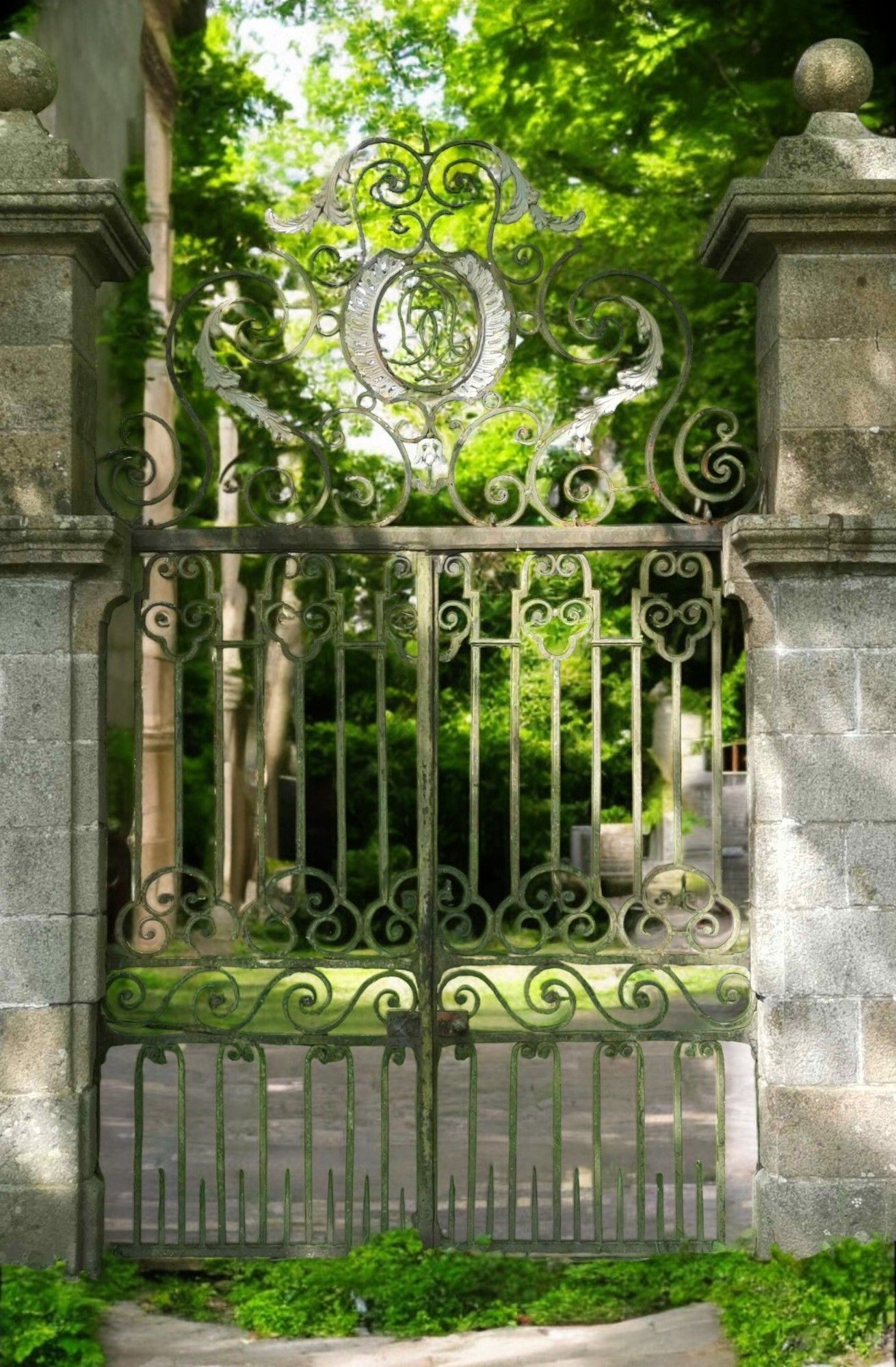 Un portail en fer forgé orné, entre des piliers de pierre, se détache sur un fond d'arbres et de verdure.