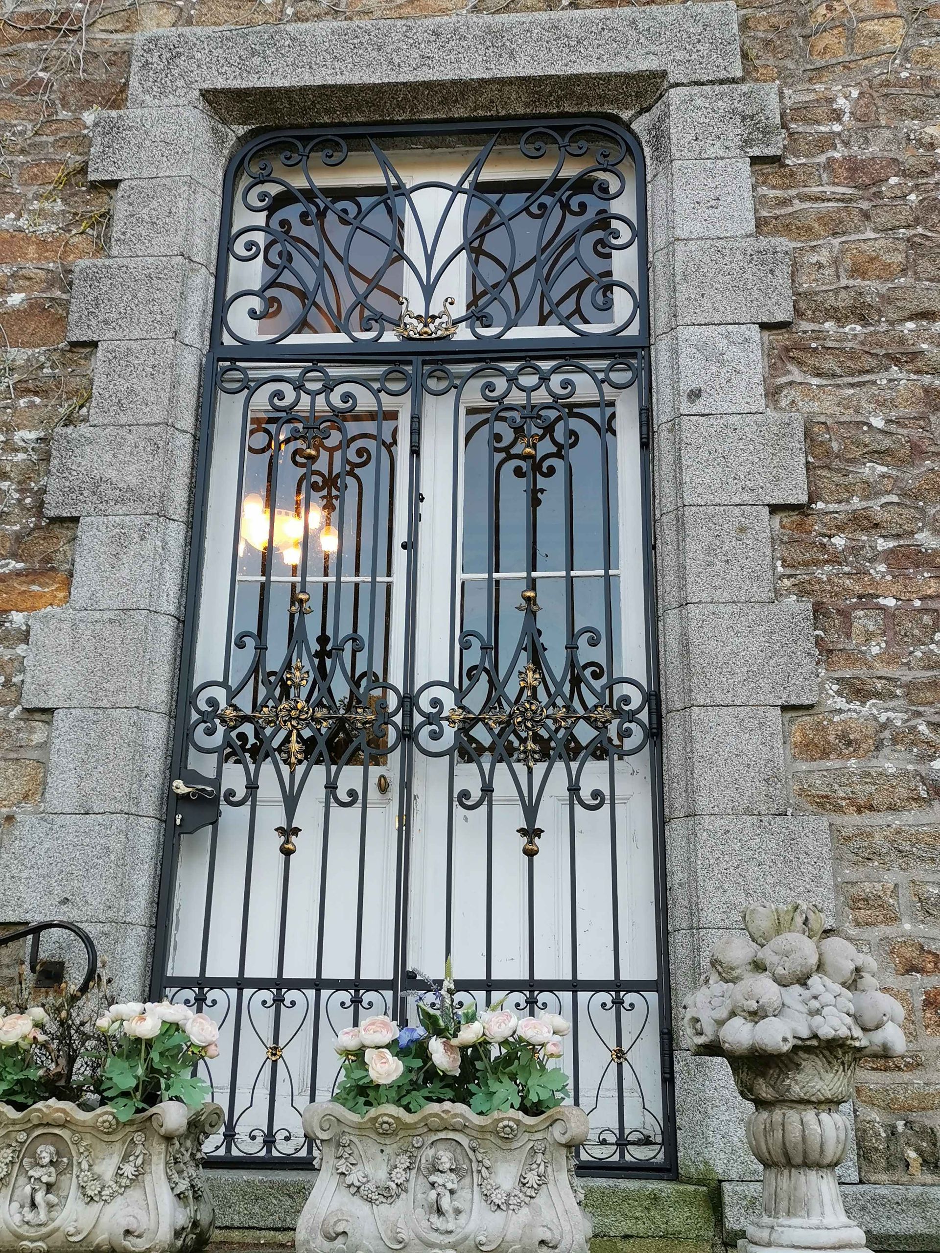 Un portail en fer noir orné et une porte blanche dans un cadre en pierre, flanqués de jardinières fleuries.