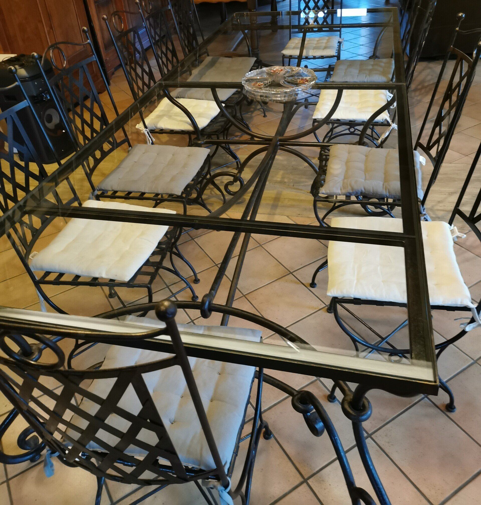 Table de jardin avec plateau en verre, chaises en fer forgé, coussins et cendrier.