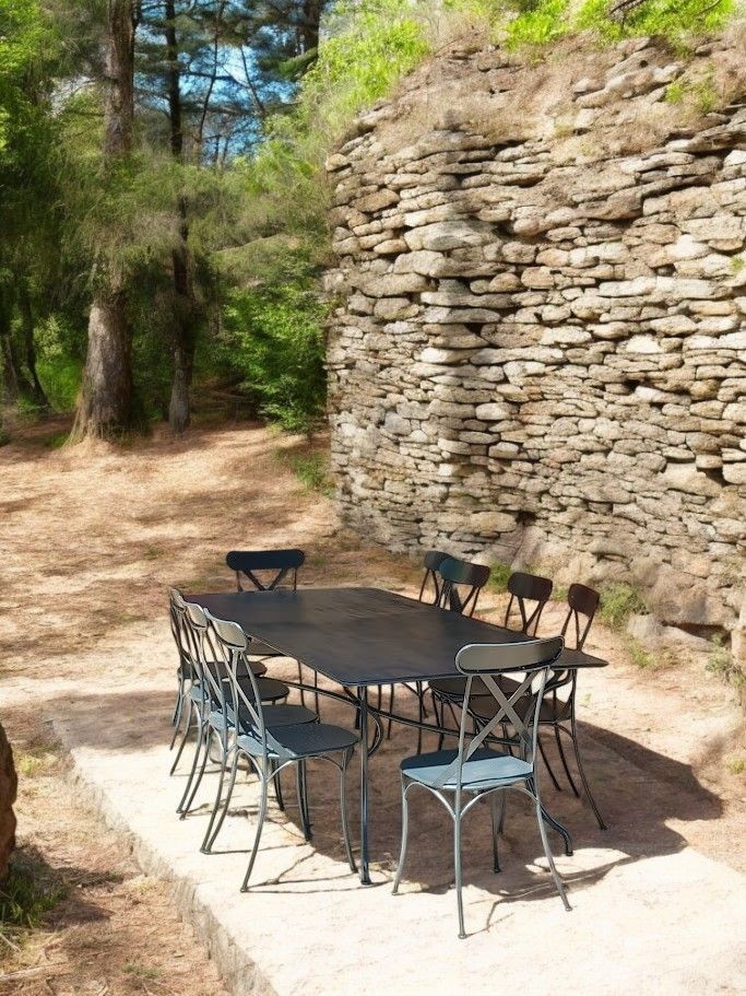 Table et chaises de jardin sur une dalle de béton près d'un mur de pierre, entourées d'arbres.
