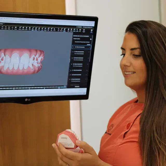 Una mujer sostiene un modelo de dientes frente a una pantalla de computadora.