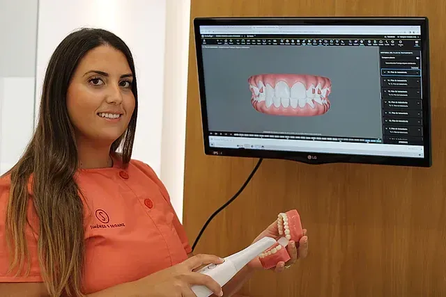 Una mujer sostiene un modelo de dientes frente a una pantalla de computadora.