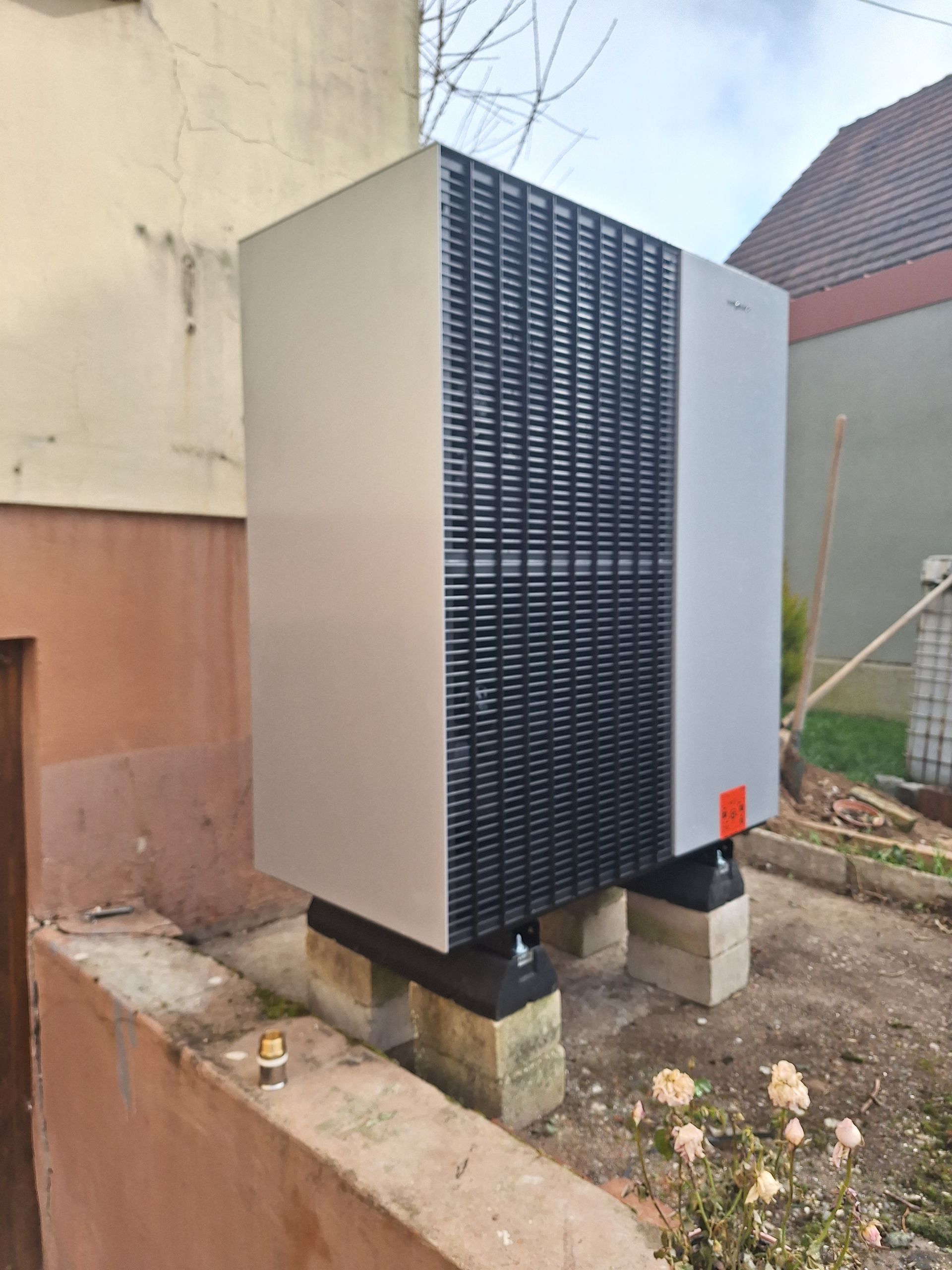 Unité de pompe à chaleur carrée de grande taille, installée sur des blocs de béton à l'extérieur, à côté d'une maison.