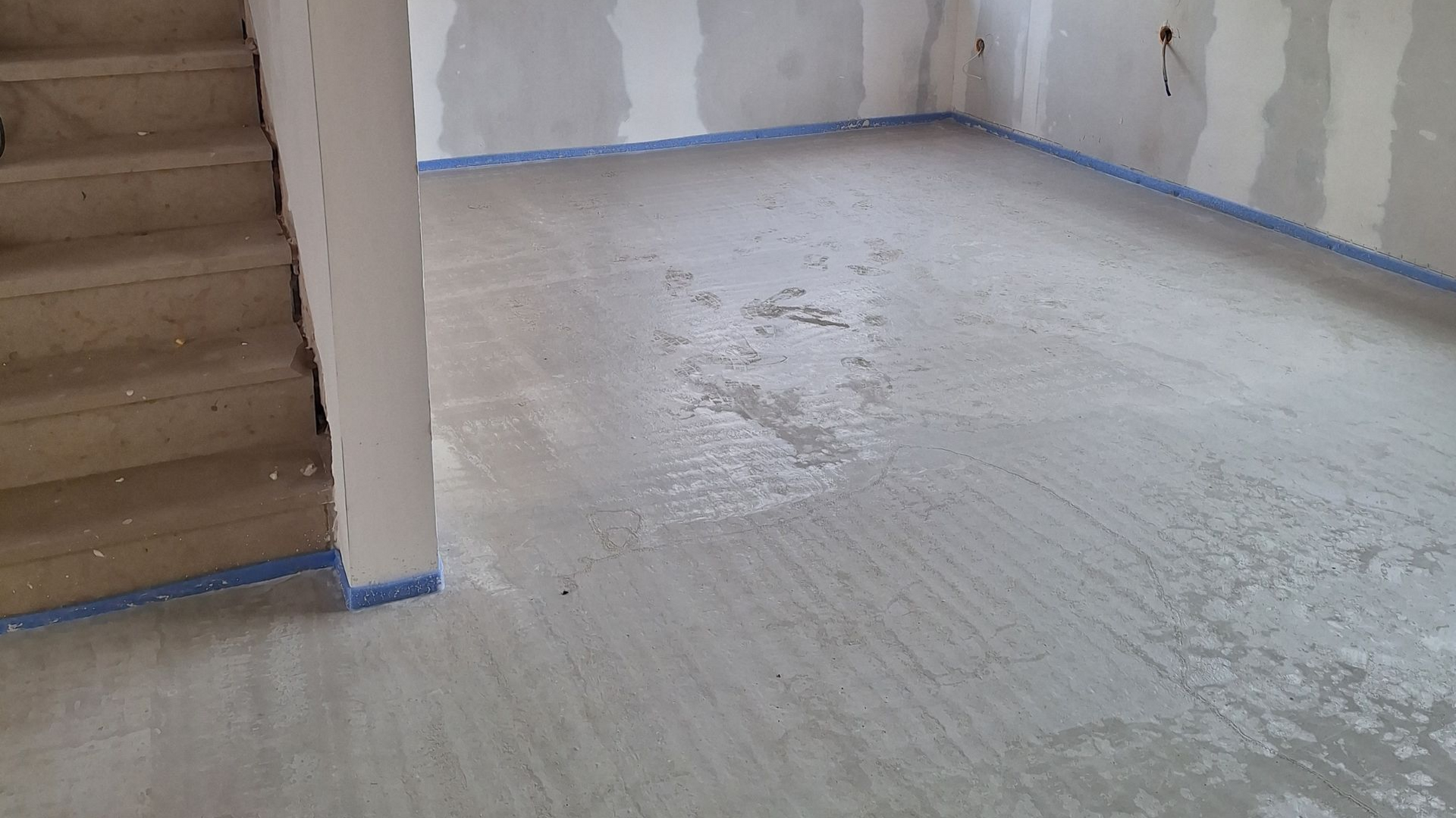 Sol en béton gris dans un sous-sol, ruban adhésif bleu le long du mur, escalier à gauche, murs non finis.